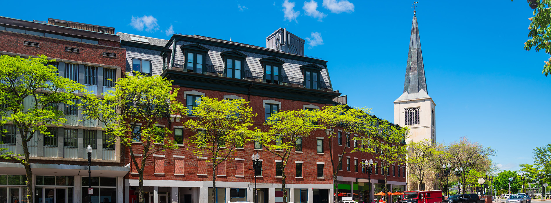 The image shows a picturesque town scene with a church steeple prominently visible against a clear blue sky, surrounded by tall buildings and trees under a bright sun.