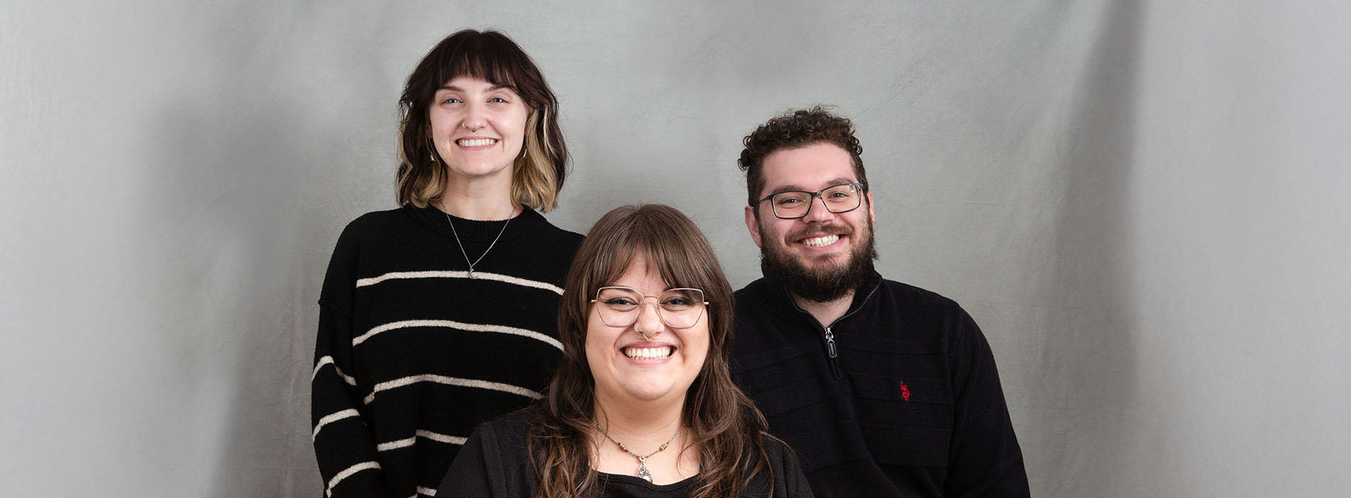 The image features three individuals posing together with smiles  two are seated and one stands behind them, all dressed in casual attire.