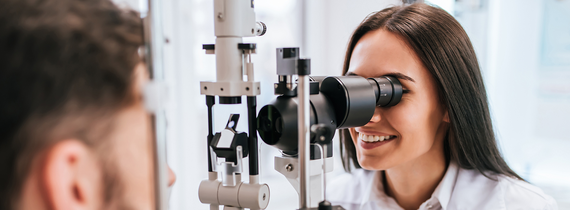 The image shows a woman wearing glasses and a white lab coat standing at an eye exam station, with a man looking into a device through her guidance.