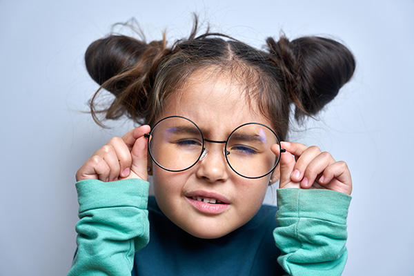 A young child with pigtails wearing glasses, making an exaggerated facial expression, with hands on face, against a white background.