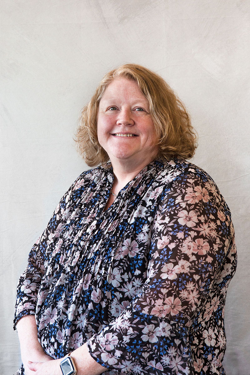 A woman standing against a white wall, smiling slightly and posing for the camera.