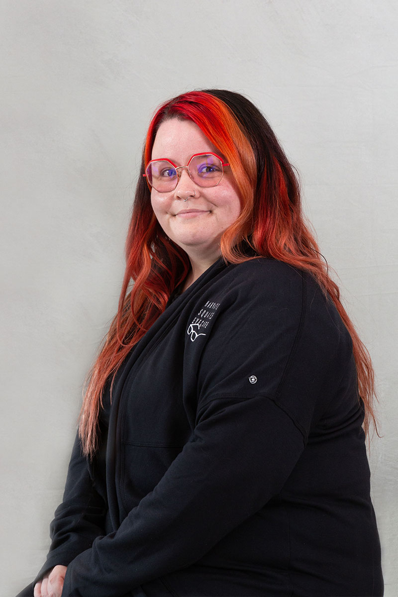 A person with colorful hair, wearing glasses, sits against a white background.