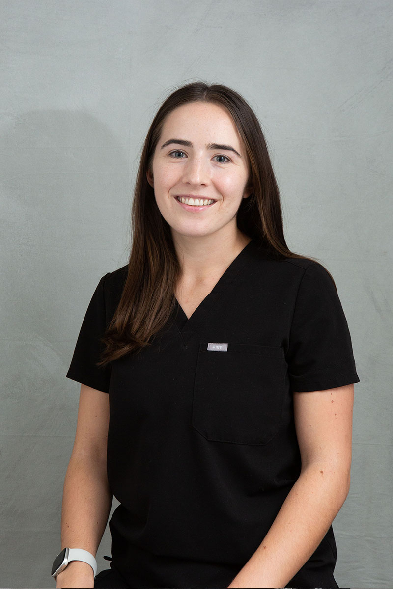 A woman wearing scrubs sits with a smile, presenting a professional appearance.
