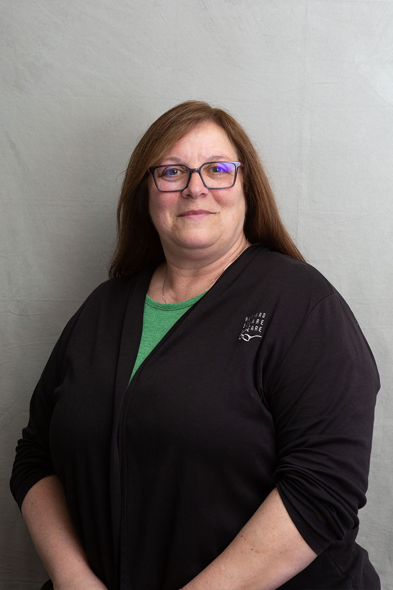 The image shows a woman standing against a plain background. She appears to be smiling and is wearing glasses, a black jacket, and has long hair.