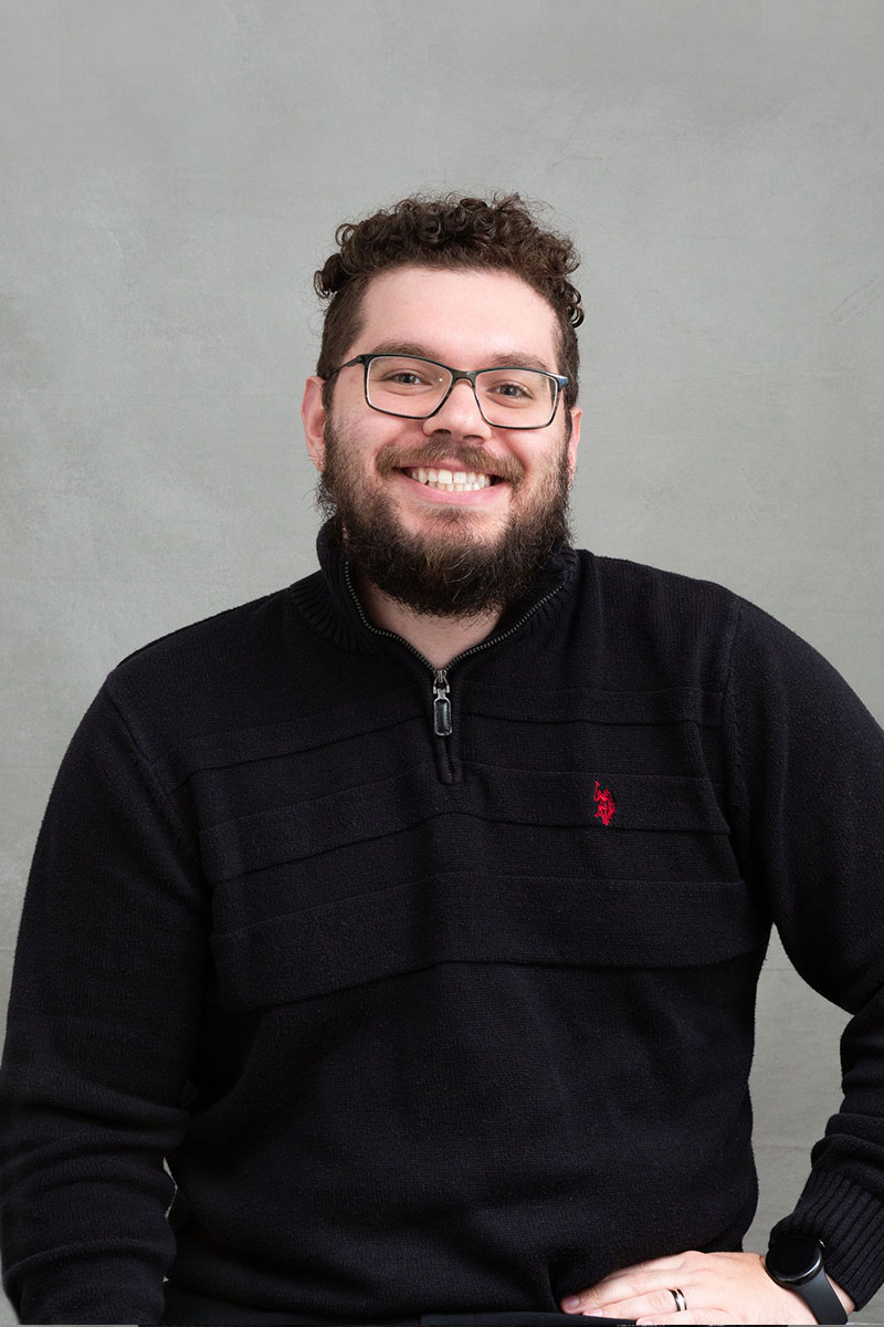 The image shows a man with glasses and a beard, wearing a dark sweater, standing with his arms crossed and smiling at the camera.