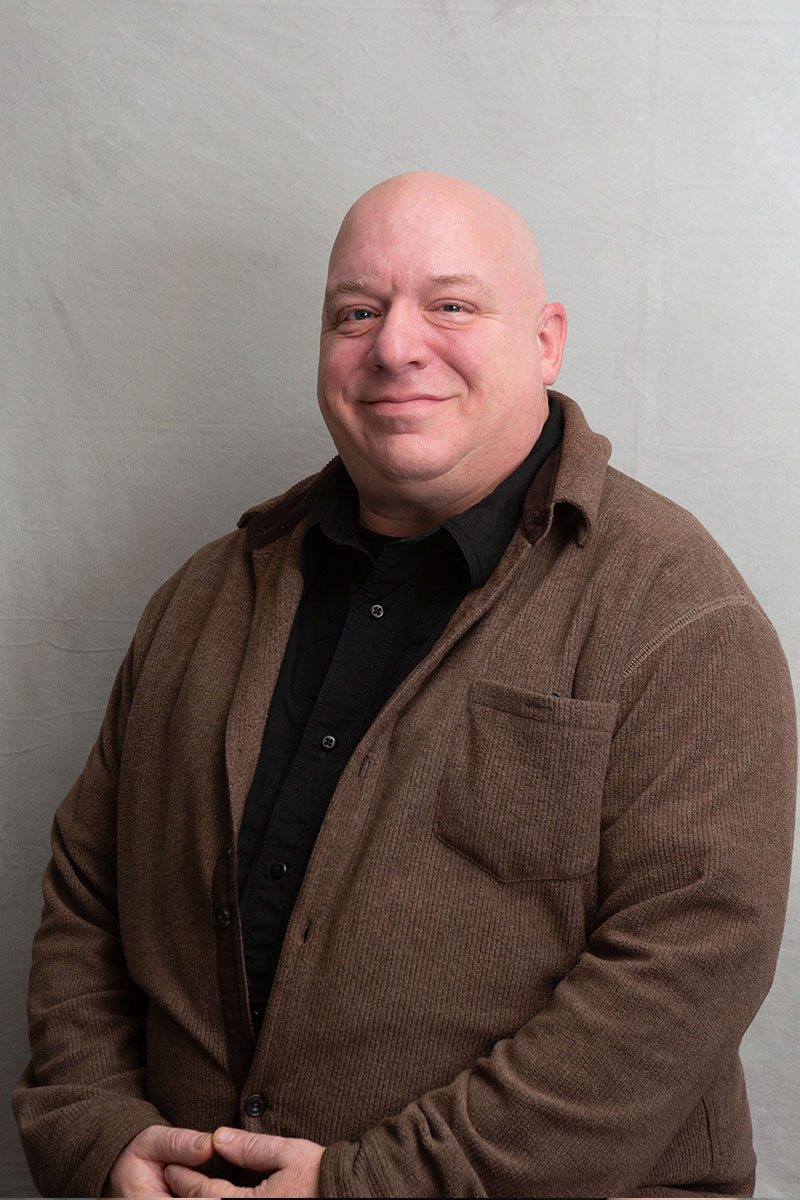 A man with a bald head stands against a white background he wears a brown jacket and has his hands clasped together in front of him.