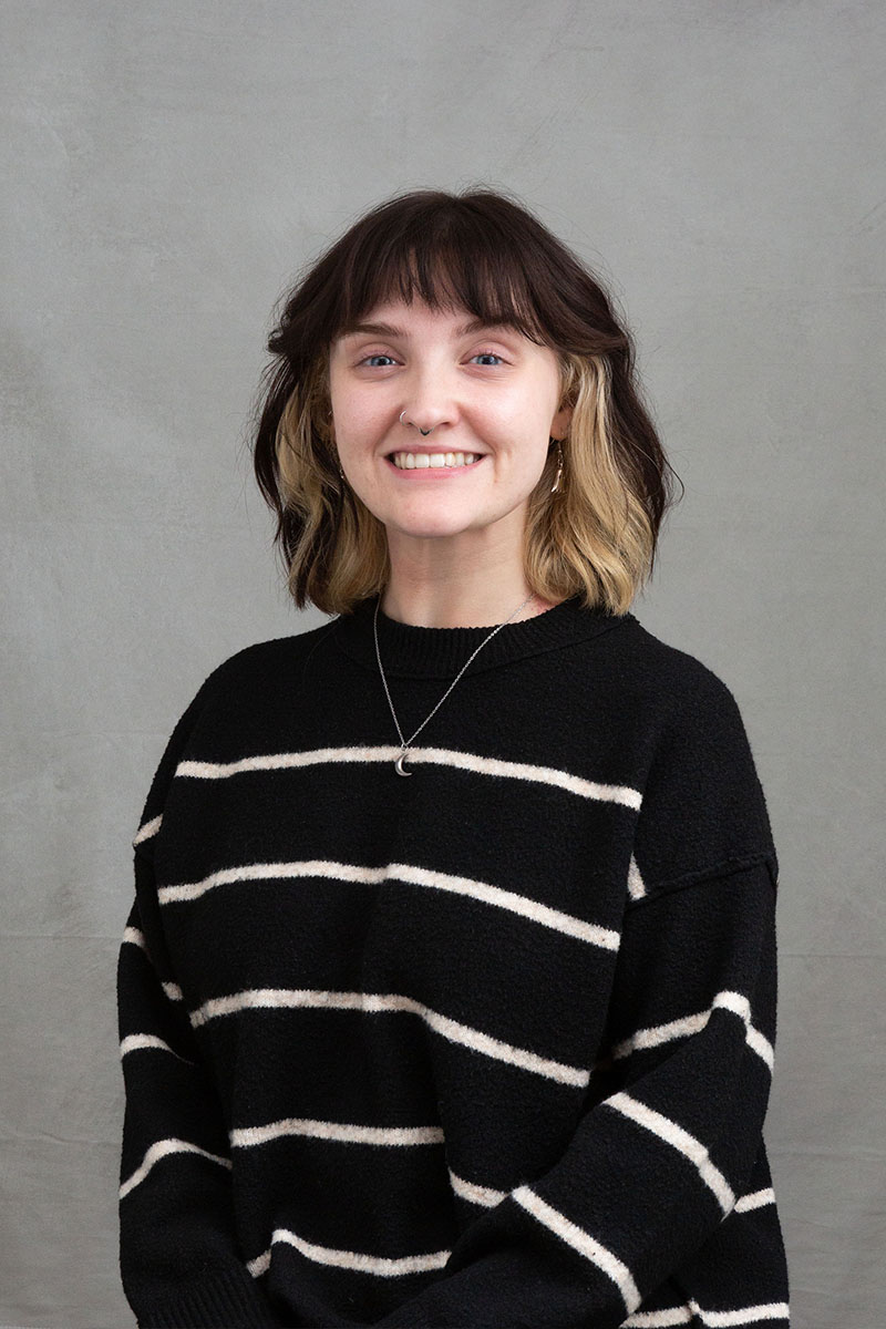 The image shows a person posing for a portrait with a neutral background. They are wearing a black and white striped sweater, have short hair, and are smiling towards the camera.