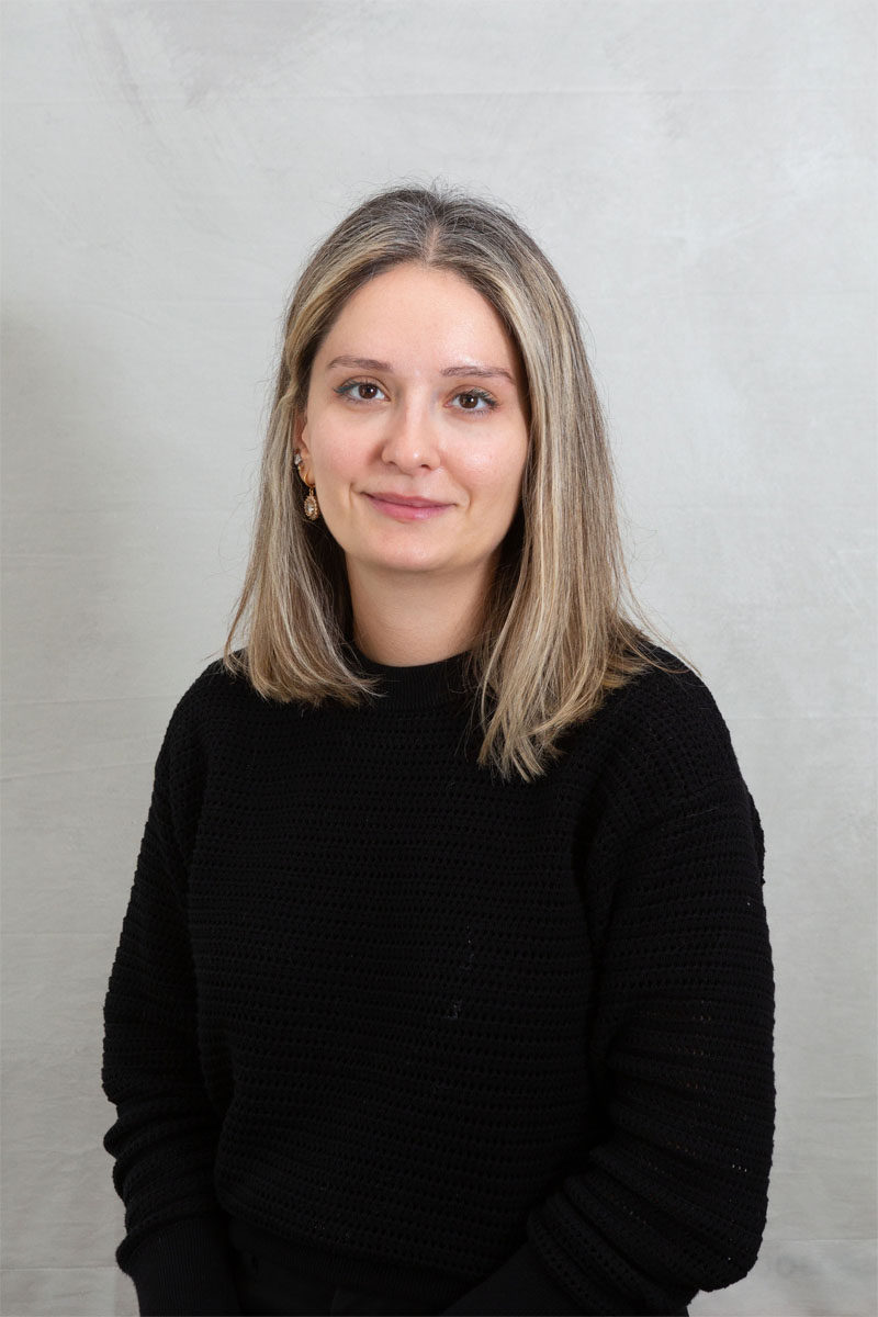 The image shows a woman posing for a portrait with a neutral background, wearing a black sweater, and smiling slightly at the camera.