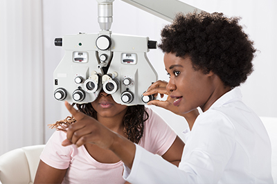A woman in a white coat is performing an eye examination on a seated individual using a large eye chart, with both wearing eye glasses.