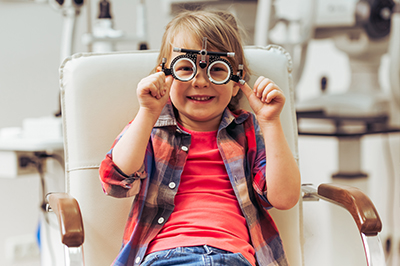 A young child wearing glasses sits in an examination chair with hands on face, smiling at camera.