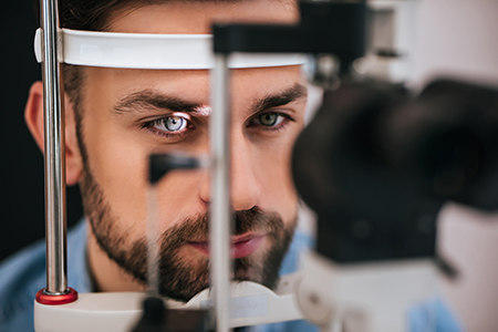 An adult man with blue eyes and facial stubble is sitting at an ophthalmologist s examination table, looking into a device with a red light, while wearing a headset-mounted eye tracker.